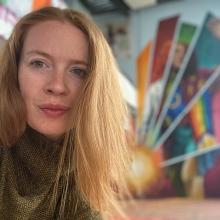 Photo of a woman with long red hair and a green jumper. Behind her is a multicoloured mural of a woman holding a lantern